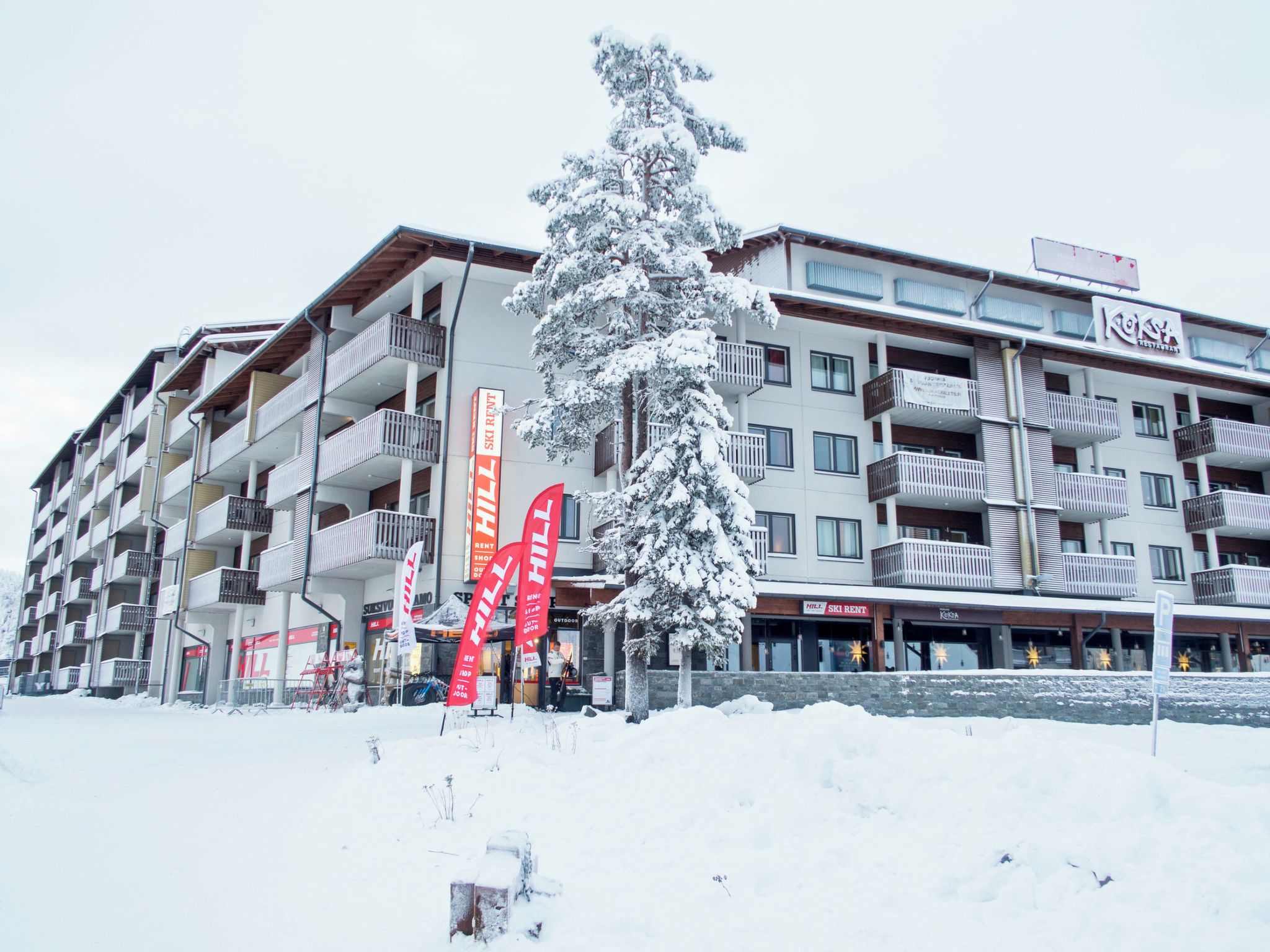 Ett hotell med flera våningar, balkonger, skyltar som visar var man kan hyra skidor och ett snötäckt träd framför.