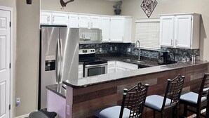 A kitchen with white cabinets, a stainless steel refrigerator, a built-in island with bar stools, and decorative wall art. - Desert Valley Stay - Mountain Views (Phoenix)
