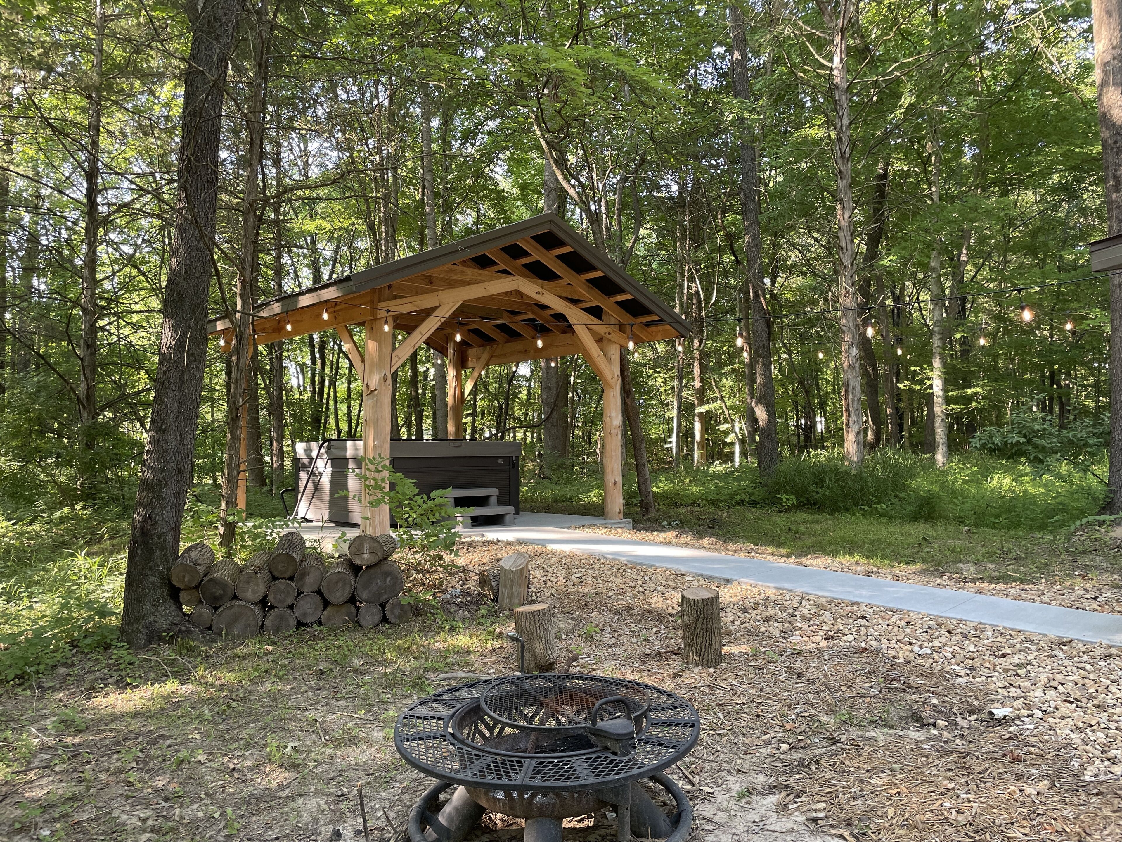 A wooden pavilion with a hot tub and a fire pit in a forested area.