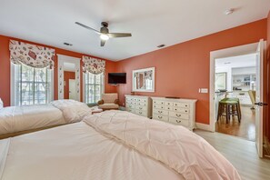 Une chambre avec deux lits, un ventilateur de plafond, une télévision et une cuisine visible à travers une porte ouverte.