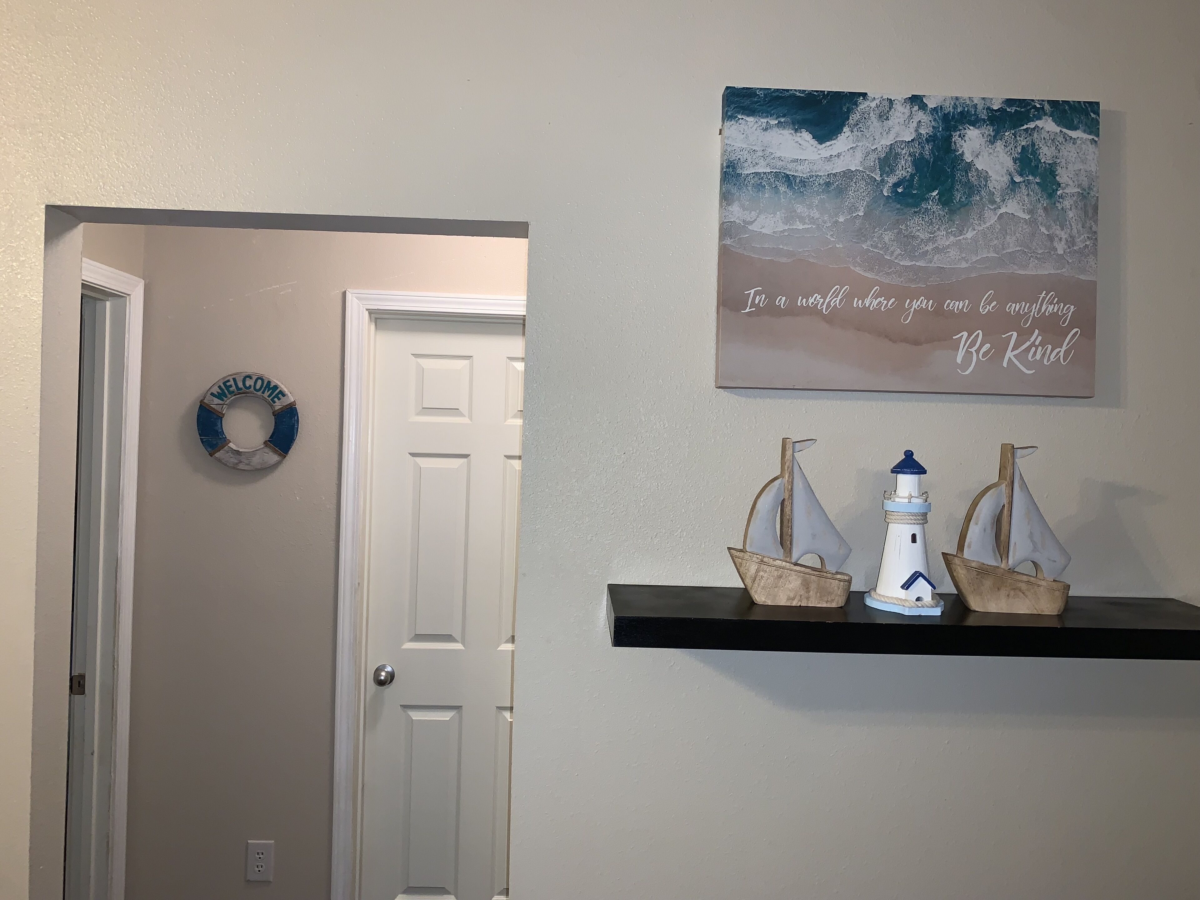 Een gang met een witte deur, een welkomstbord, een wanddecoratie met een strandgezicht en een citaat, en een plankje met twee kleine bootjes en een vuurtoren.