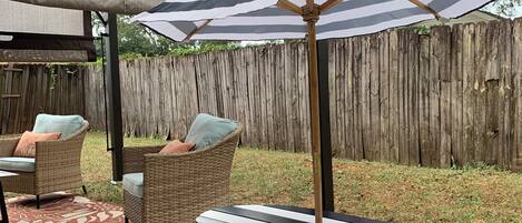 A wooden picnic table with benches, a striped umbrella, wicker chairs, and a covered area.