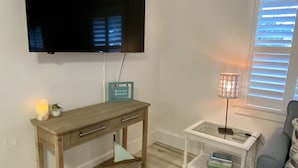 A living room with a wooden desk, a television mounted on the wall, a small table with a lamp, and a blue sofa. - Chic cottage, Vamo area, close to beaches, pet friendly, kayaks, large backyard  (Sarasota)