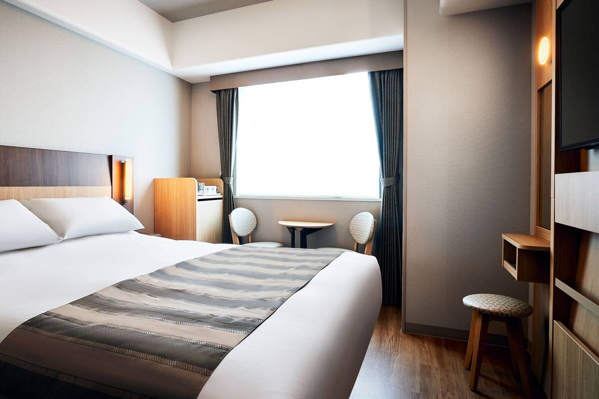 A hotel room with a large bed, a desk, a chair, a TV, and a window with curtains.