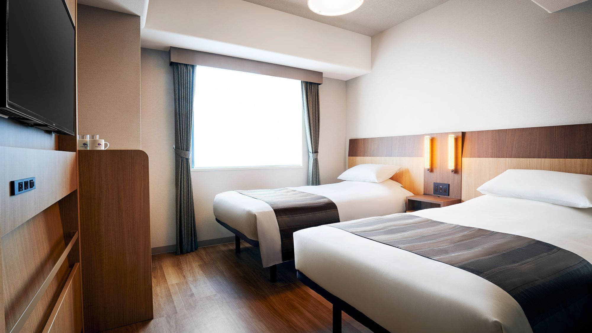 A hotel room with two beds, a TV, a bedside table with lamps, and a window with curtains.