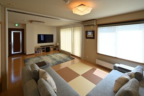 Ein geräumiges Wohnzimmer mit einer großen Couch, einem Flachbildfernseher, einem Bücherregal und einem großen Fenster mit Jalousien.