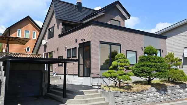 A modern two-story house with a garage, a covered porch, and a well-maintained garden with small trees. - Casi 15 (Furano)