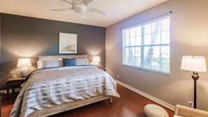 Una camera da letto con un letto, due abat-jour, una ventola a soffitto, una finestra con persiane e un'opera d'arte appesa al muro.