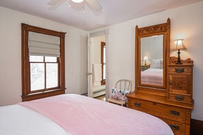 Ein Schlafzimmer mit einem Bett, einer hölzernen Kommode mit Spiegel, einem Sessel und einem Fenster mit weißem Rollo.