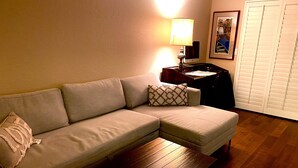 Una sala de estar con un sofá, una mesa de madera, una lámpara, un escritorio con una computadora y un cuadro en la pared.