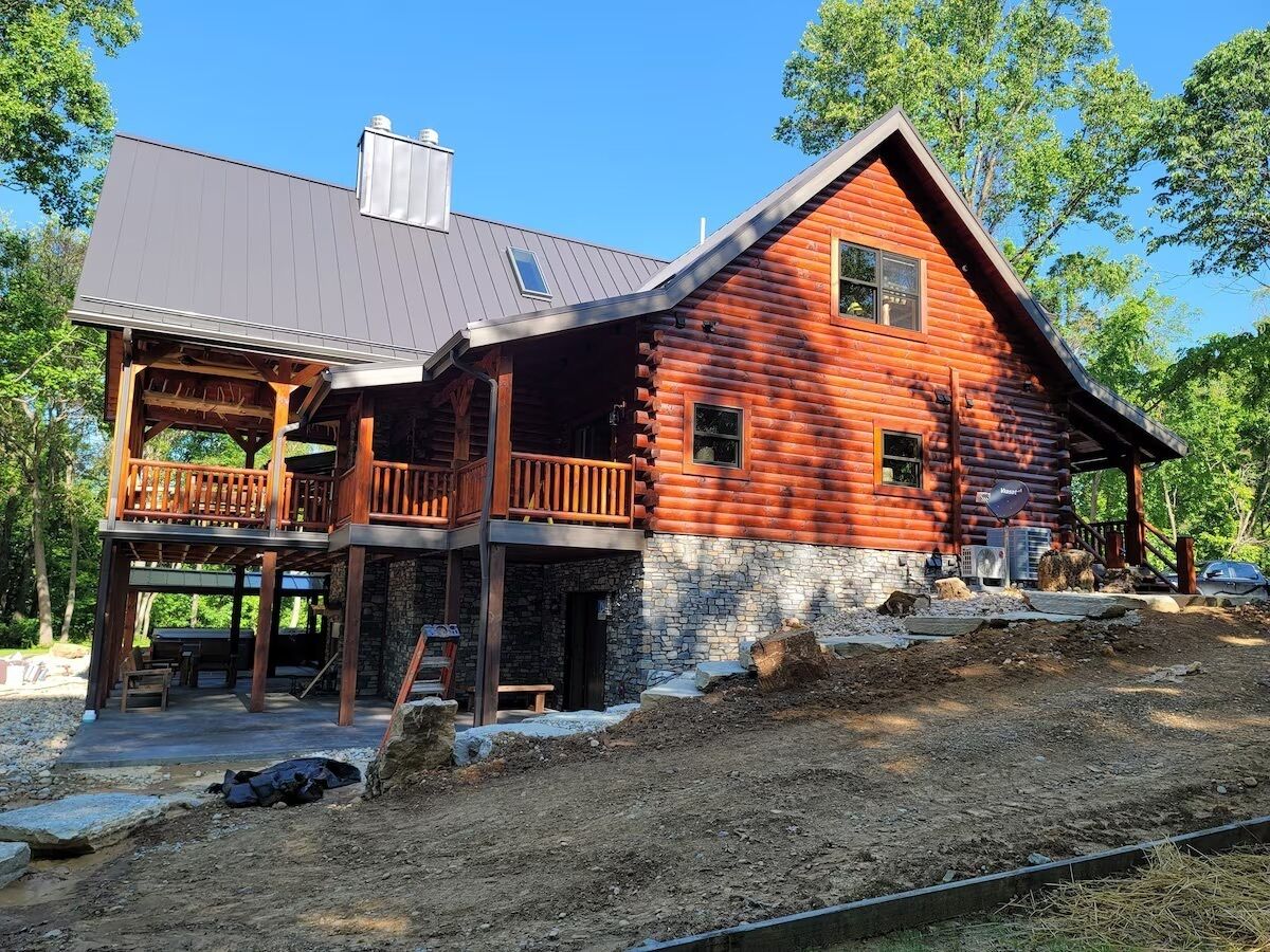 A two-story log cabin with a balcony, stone foundation, and a metal roof.