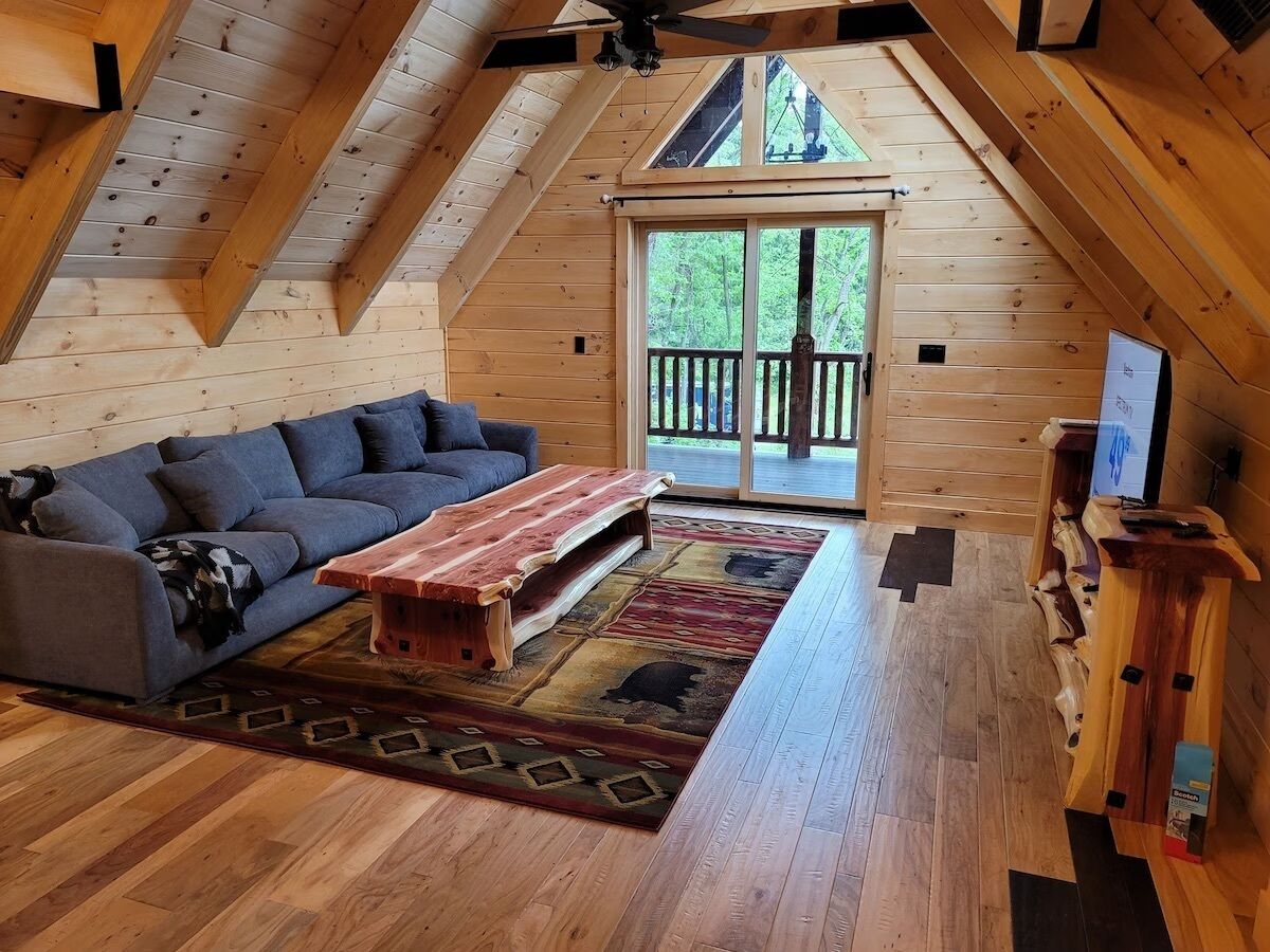 A cozy loft-style room with a wooden ceiling, a gray sofa, a wooden coffee table, and a large window with a view of trees.