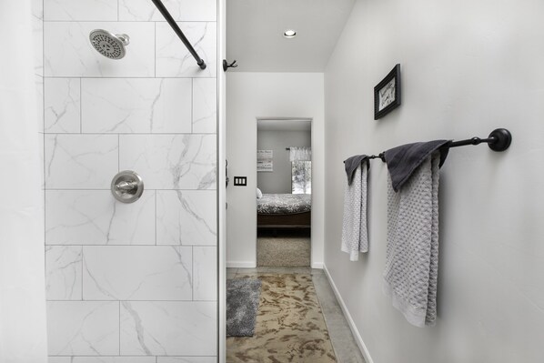 A bathroom with white marble tiles, a shower with a handheld showerhead, a towel rack with towels, and a visible door leading to a bedroom.