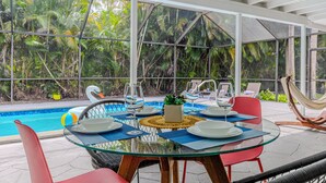 Un espace repas entièrement vitré, avec vue sur la piscine, comprenant une table ronde pour deux personnes, des chaises rouges et un hamac.