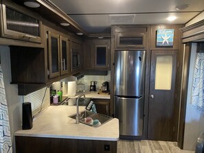 A compact kitchen with dark wood cabinets, a stainless steel refrigerator, and a "Welcome" sign above the pantry door.