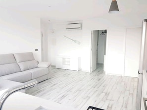 Un soggiorno moderno con un divano grigio, un tavolino da caffè bianco e un pavimento in legno. Si vede una porta bianca che conduce in un'altra stanza e una lampada a sospensione.