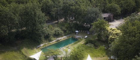 An aerial view of a secluded outdoor area with a swimming pool, surrounded by dense forest, and featuring two white tents.