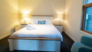 A neatly made bed with white linens, two bedside lamps, a window with blinds, and a blue chair. - Snow Dayz - Dinner Plain (Dinner Plain)