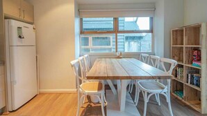 A dining area with a wooden table and white chairs, a refrigerator, and a bookshelf. - Snow Dayz - Dinner Plain (Dinner Plain)