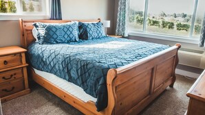A wooden bed with a quilted blue and white bedding, a nightstand with drawers, a bedside lamp, and a window with a view of greenery. - Beautiful 2 bedroom home with amazing views (Gaston)