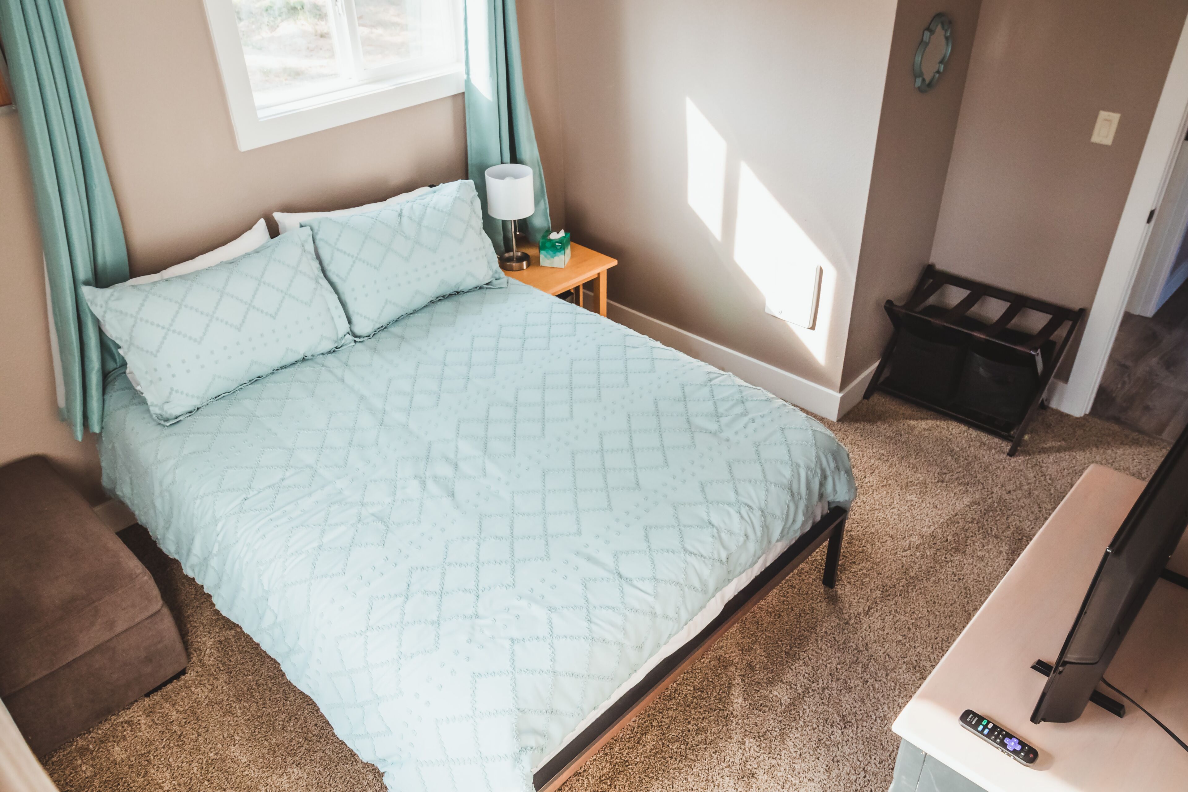 A bedroom with a bed, a nightstand, a lamp, a remote control, a TV, and a small trash bin.