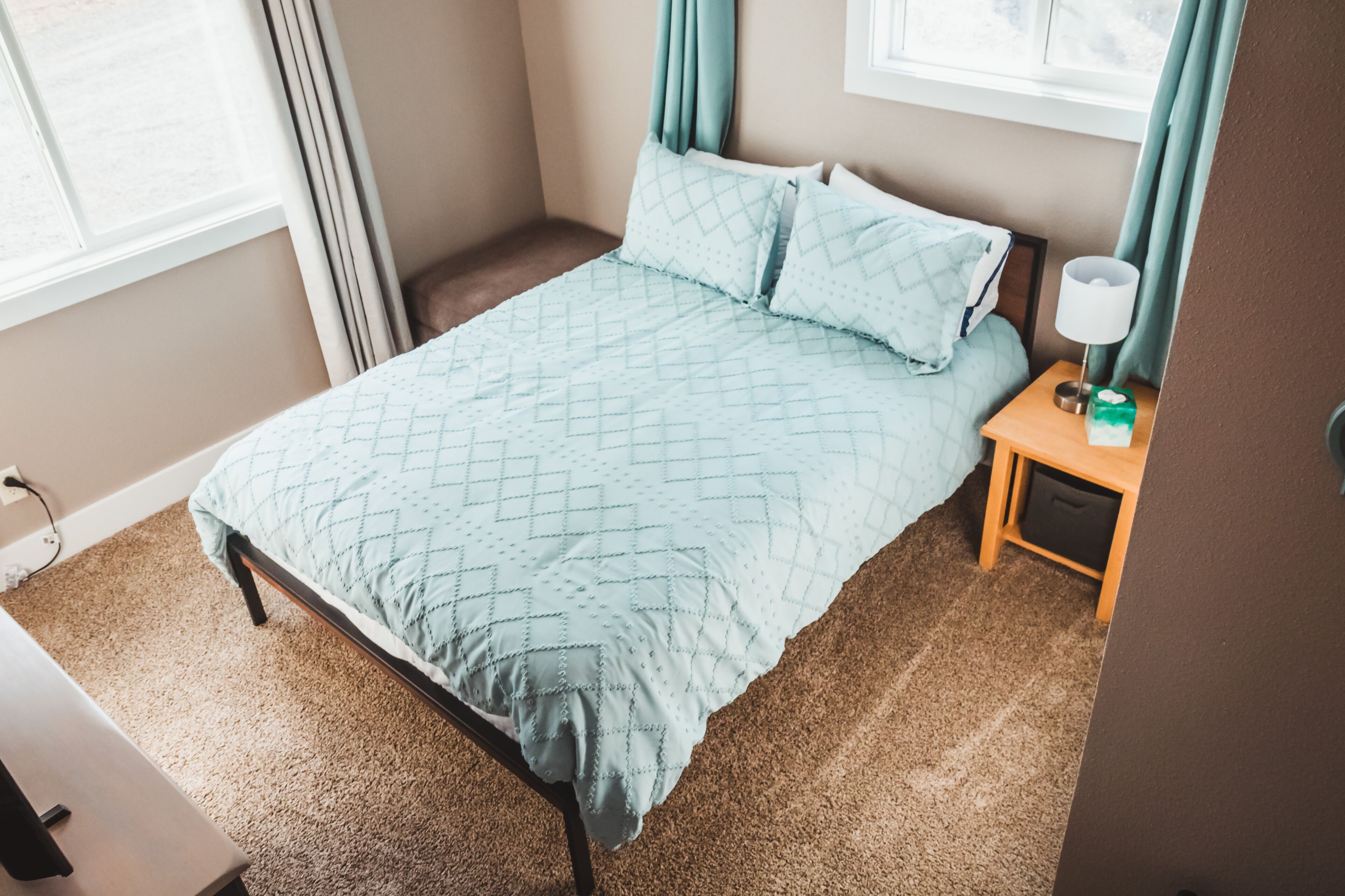 Un dormitorio con cama, mesita de noche, lámpara y ventana con cortinas.