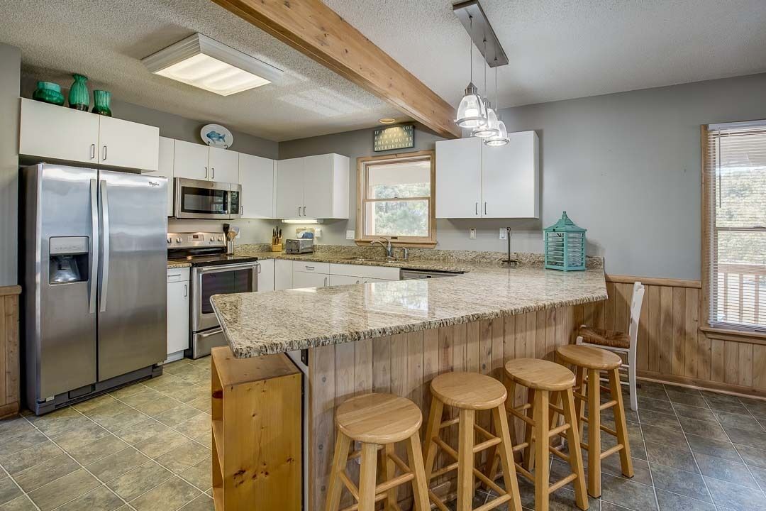 A kitchen with a large island, wooden stools, stainless steel appliances, and a window with blinds.