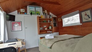 Una habitación acogedora con techo de madera, una cama con manta color beige, un escritorio con silla, una estantería y una ventana con cortinas.
