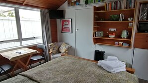 Una habitación acogedora con una cama, un escritorio y una silla. Hay estantes con libros y utensilios de cocina.