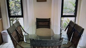 Un comedor con una mesa de vidrio rodeada de sillas de mimbre, ventanas amplias y una lámpara colgante.