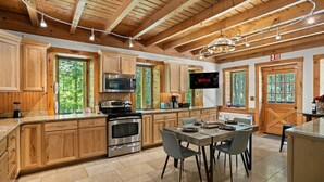 Una cocina moderna con armarios de madera, electrodomésticos de acero inoxidable, una zona de comedor con mesa y sillas, y un amplio ventanal con vistas al exterior.