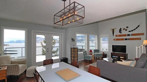 Un salón amplio con una mesa blanca grande, un sofá gris y un televisor. Hay ventanas grandes que ofrecen vistas al mar.