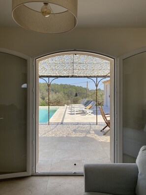 A room with a sliding glass door leading to a pool area with sun loungers and a pergola.