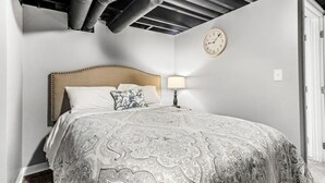 Ein Schlafzimmer mit einem großen Bett, einer Kopfteil, einer Nachttischlampe, einer Wanduhr und freiliegenden Lüftungsrohren.