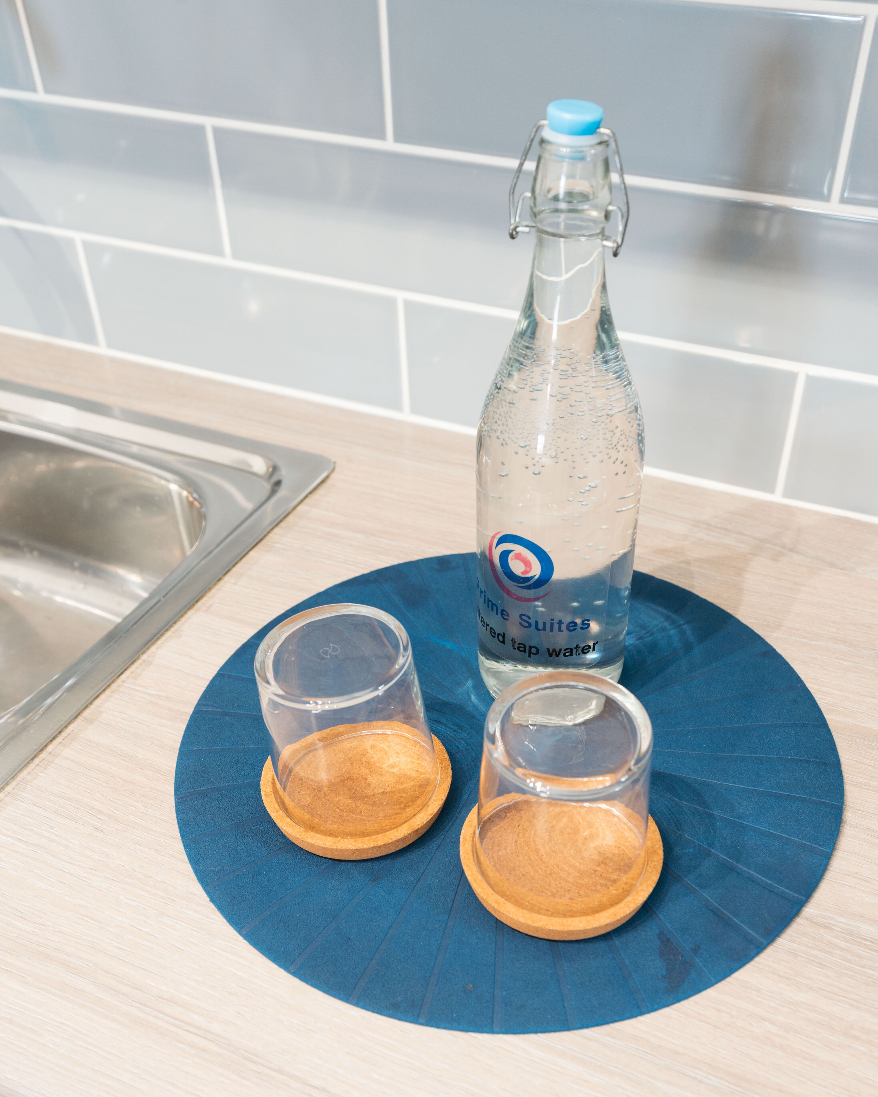 Un comptoir de cuisine avec un tapis bleu, une bouteille d’eau et deux verres posés sur des sous-verres en bois.