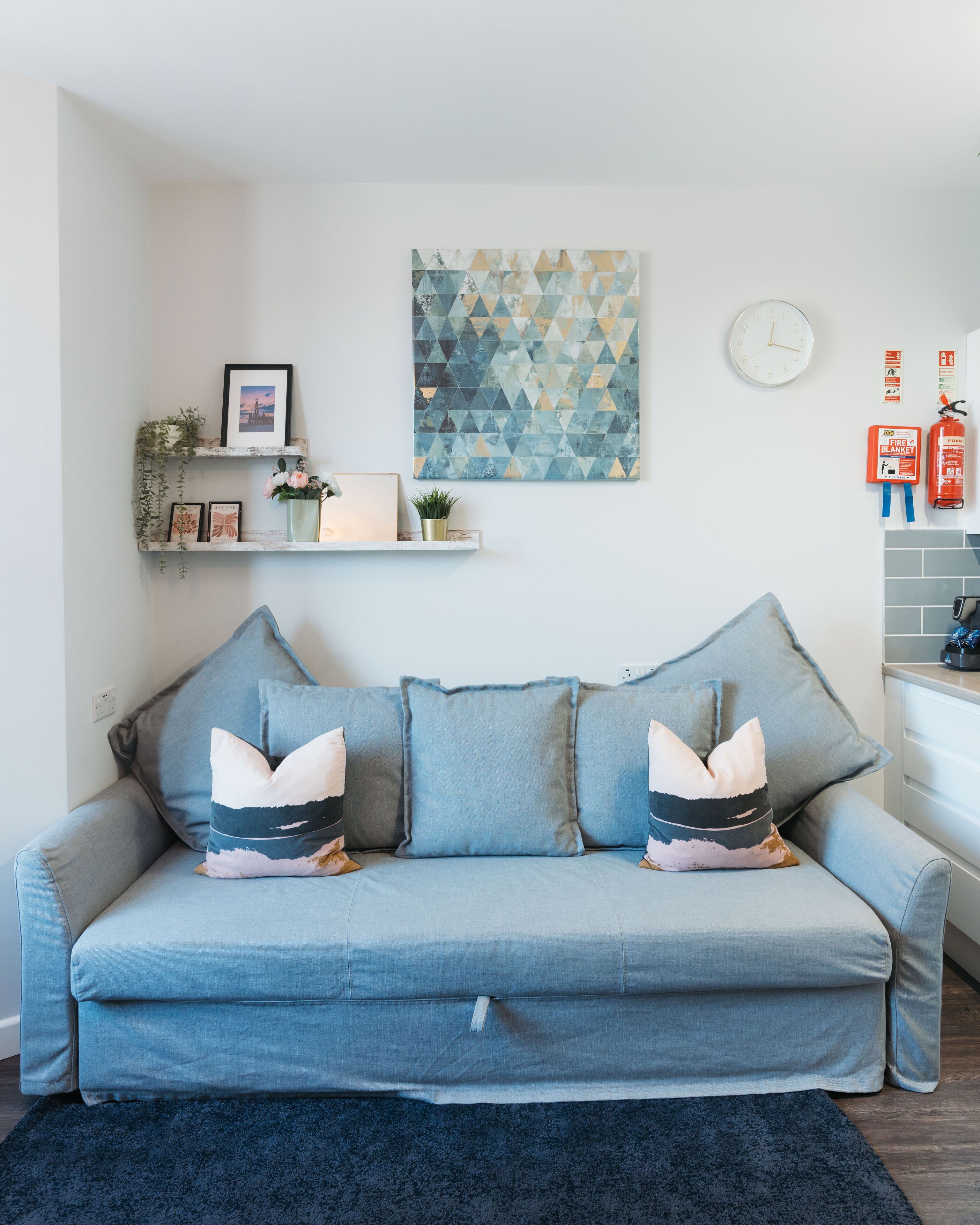 A living room with a blue sofa, a geometric wall art piece, a clock, and a fire extinguisher.