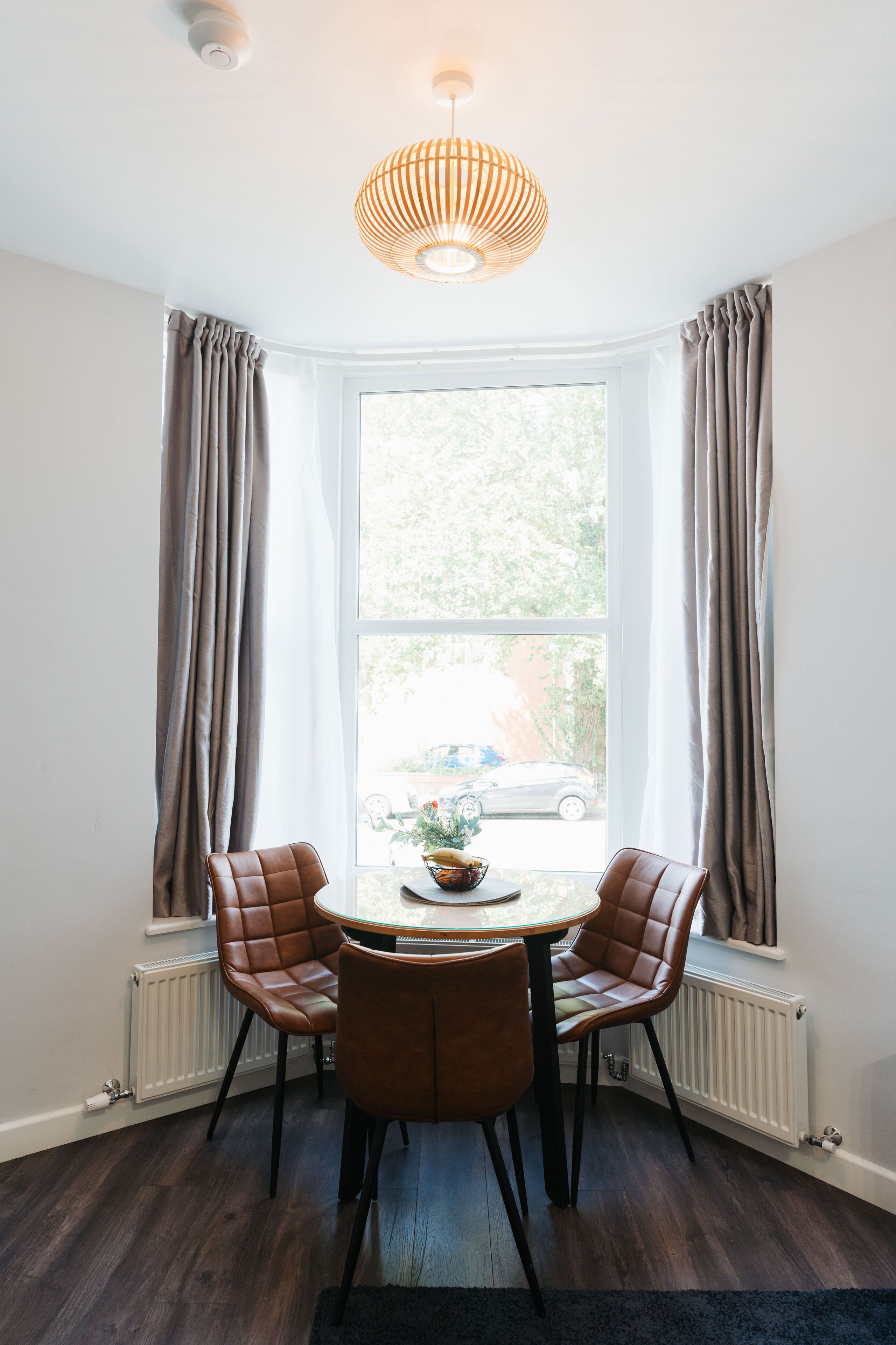 A dining area with a round table, four chairs, a hanging light fixture, and a view through a large window.
