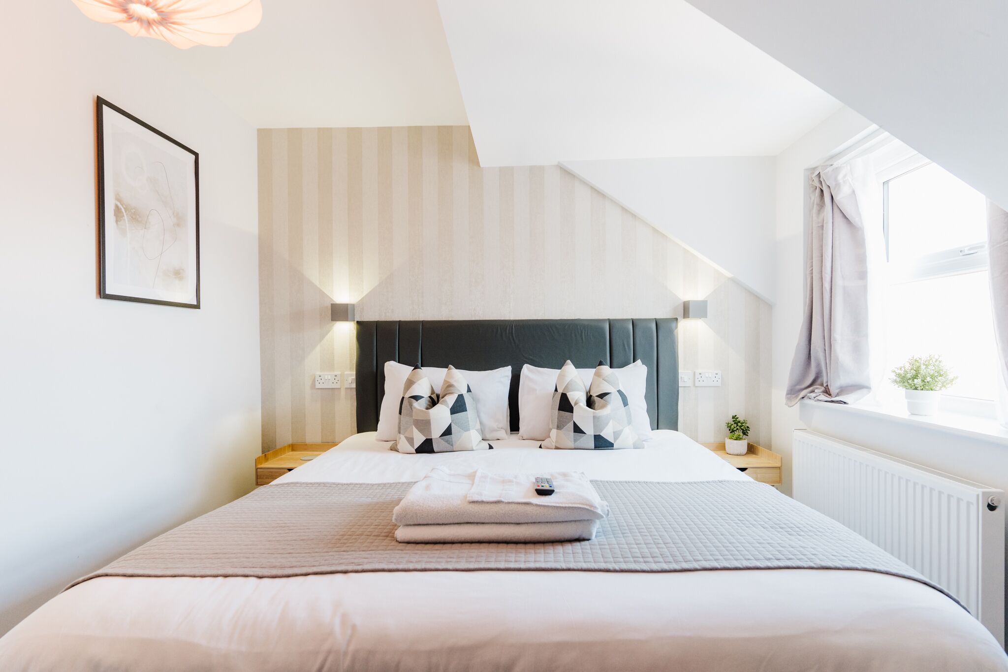 Ein ordentlich bezogenes Bett mit einem gefalteten Handtuch und Fernbedienung, ein Nachttisch mit Lampe und ein Fenster mit Vorhängen.