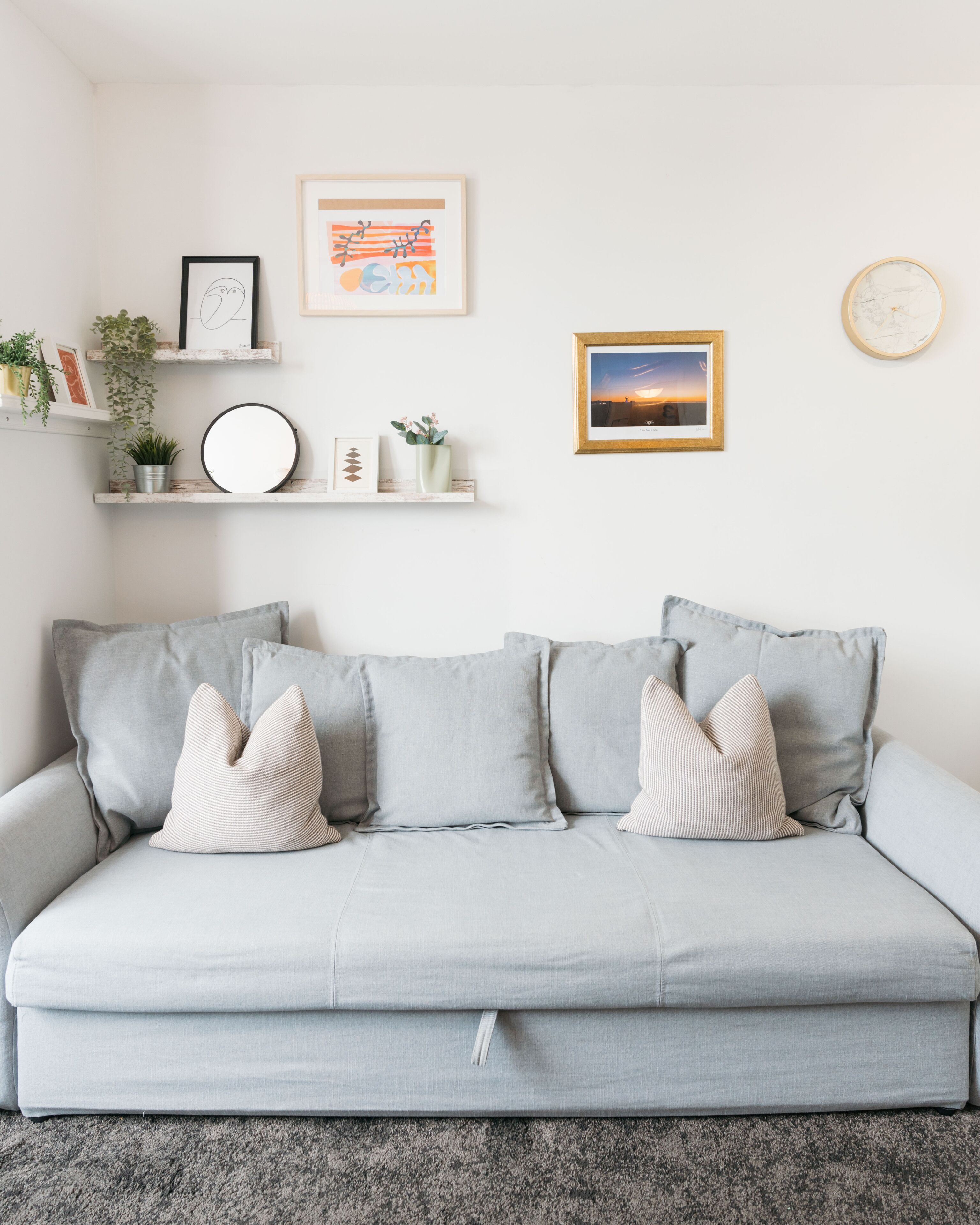 A light grey sofa with three pillows, a round mirror, and two framed artworks on a white wall.