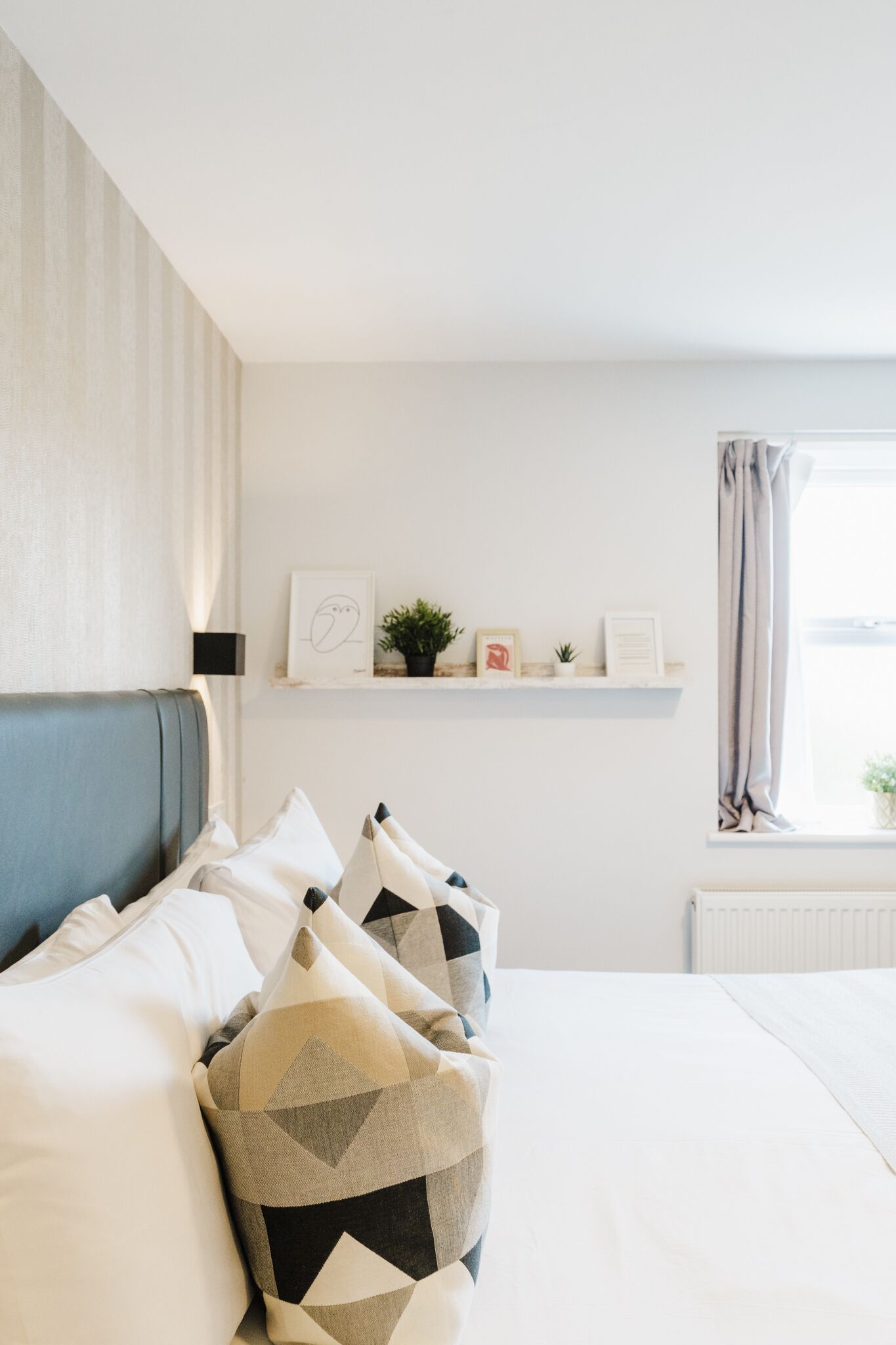 A neatly arranged bedroom with a bed, pillows, a shelf with decorative items, and a window with curtains.