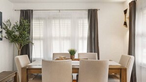 A dining area with a wooden table, four chairs, a potted plant, and sheer curtains.