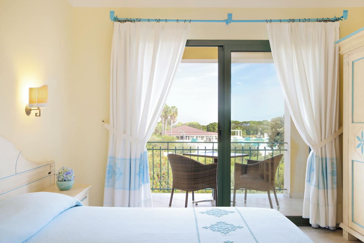 A hotel room with a bed, a small table with a flower vase, a wicker chair, and a balcony with a view of the outside.