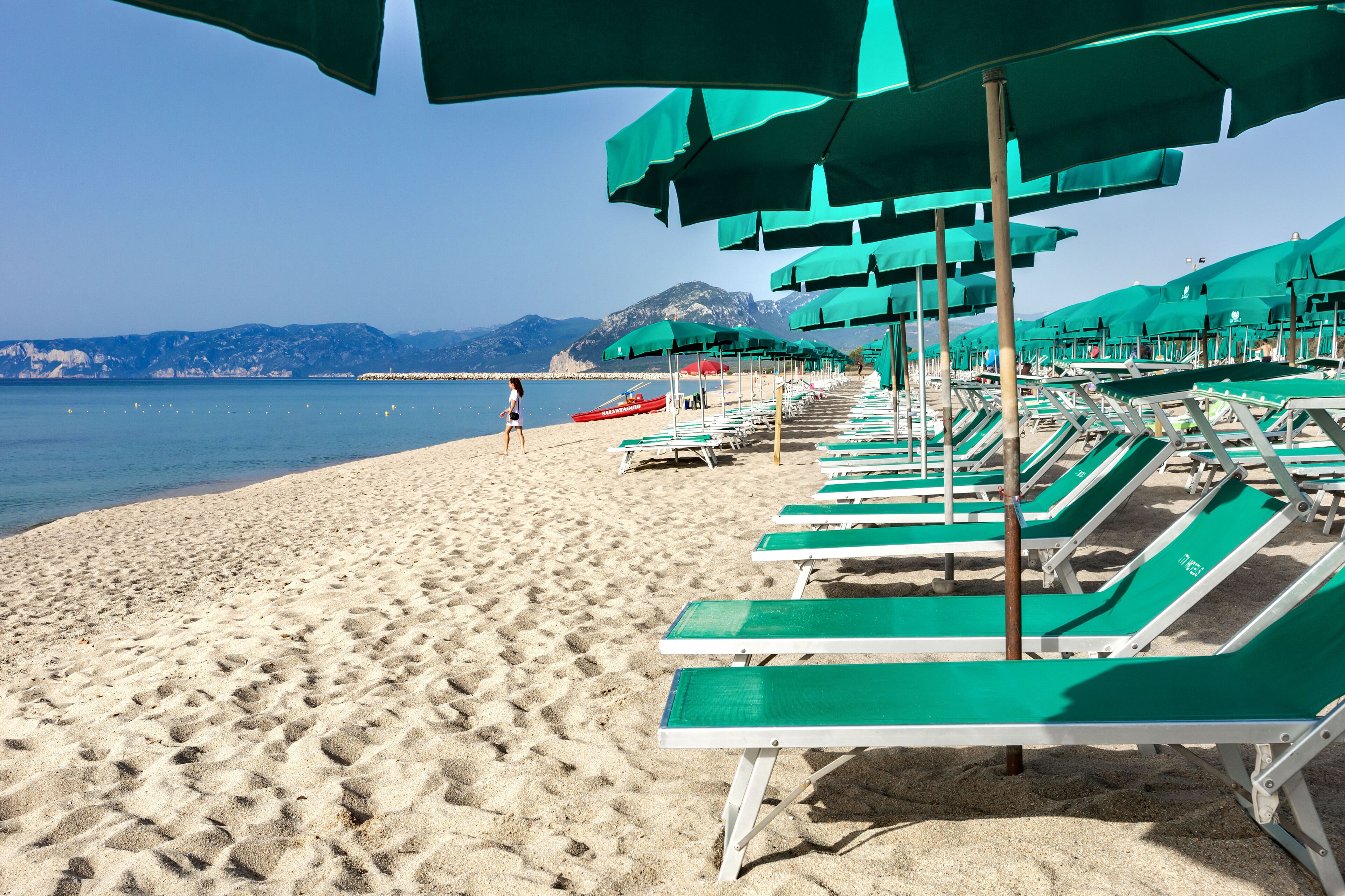 A beach with rows of green lounge chairs and umbrellas, a sandy shore, and a mountain in the background.