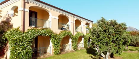 A two-story building with a balcony on each floor, surrounded by greenery and a clear sky.