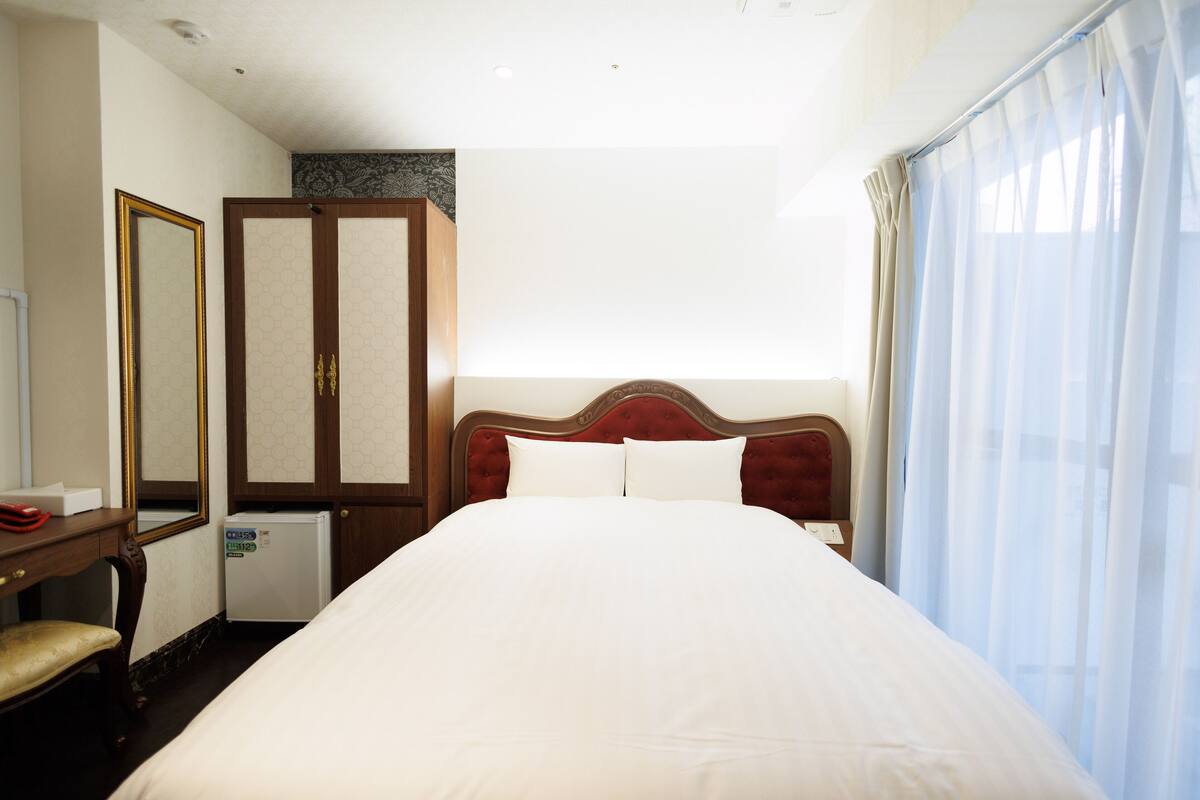 A hotel room with a large bed, a wardrobe, a desk with a chair, a mirror, and a window with curtains.