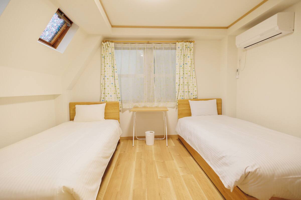 A room with two single beds, a small table, a window with curtains, and an air conditioning unit.