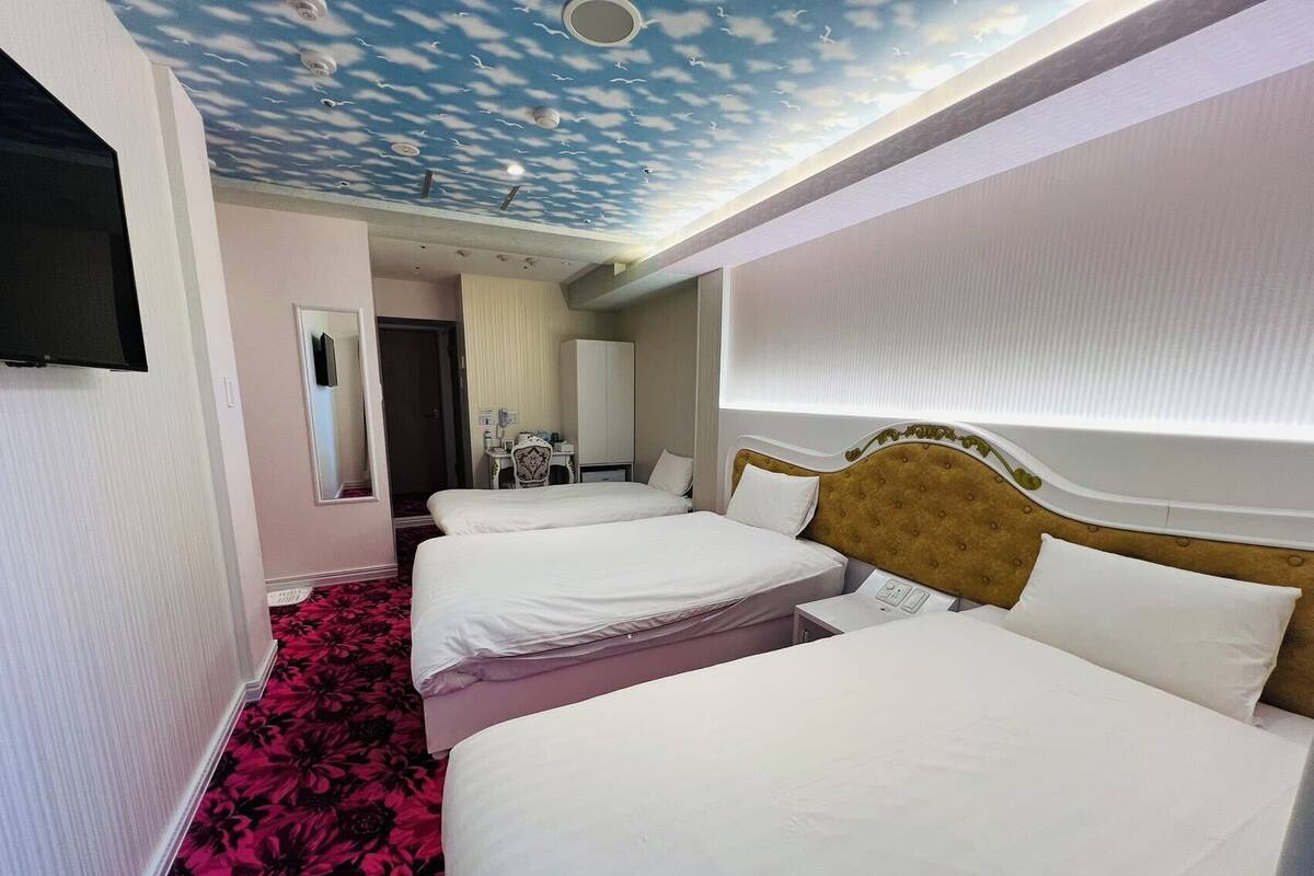 A hotel room with three beds, a TV mounted on the wall, a ceiling with a cloud design, and a floral carpet.