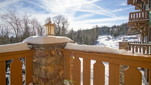 Un balcón cubierto de nieve con un muro de piedra y barandilla de madera, con vistas a un paisaje nevado y una zona boscosa.
