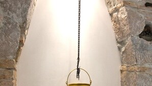 Un intérieur rustique avec un mur en pierre, un pot en laiton suspendu, un tableau décoratif représentant un feu, un panier en osier et une petite table sur laquelle se trouvent un bol et une clé.