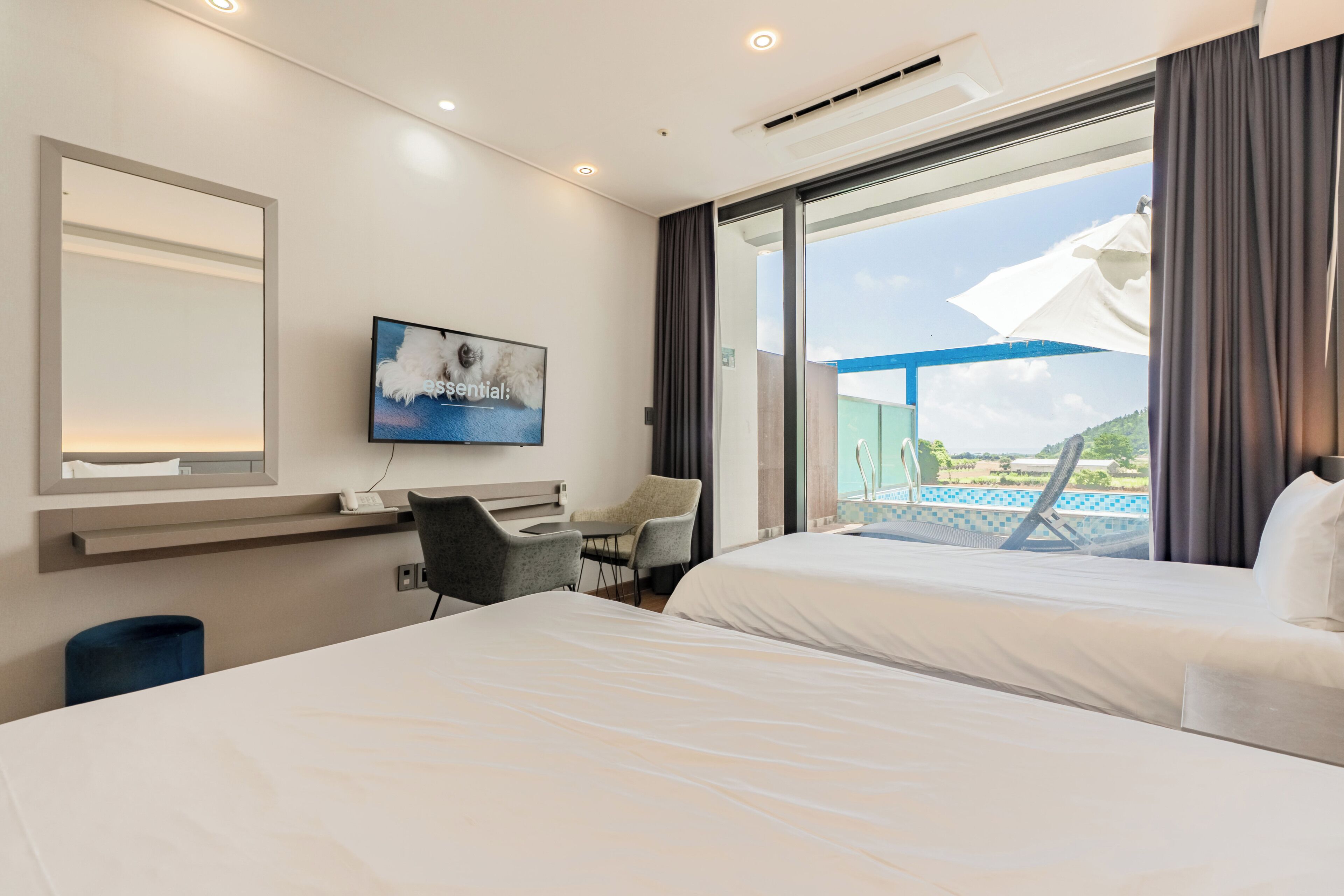A hotel room with two beds, a desk, a chair, a TV, and a large window with a view of a pool and mountains.